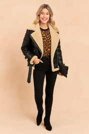 Black shearling aviator jacket with oversized beige faux-fur lining, wide lapels, belted cuffs with buckles, and zipper details, styled over a leopard-print sweater. Paired with fitted black skinny pants and black ankle boots for a bold, fashion-forward look.