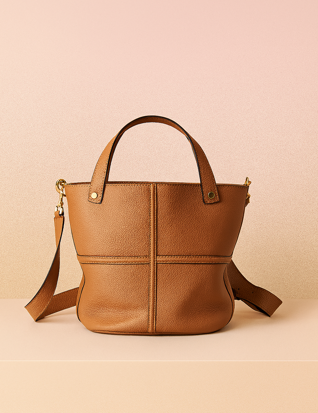 Medium-sized tan pebbled leather tote bag with structured cross-stitch detailing, gold-tone rivets, top handles, and a detachable shoulder strap, presented on a neutral display platform against a warm gradient background.