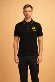 Black short-sleeve polo shirt with gold embroidered crest logo, contrast gold collar piping, three-button placket, and matching black tailored trousers with a clean straight-leg fit.