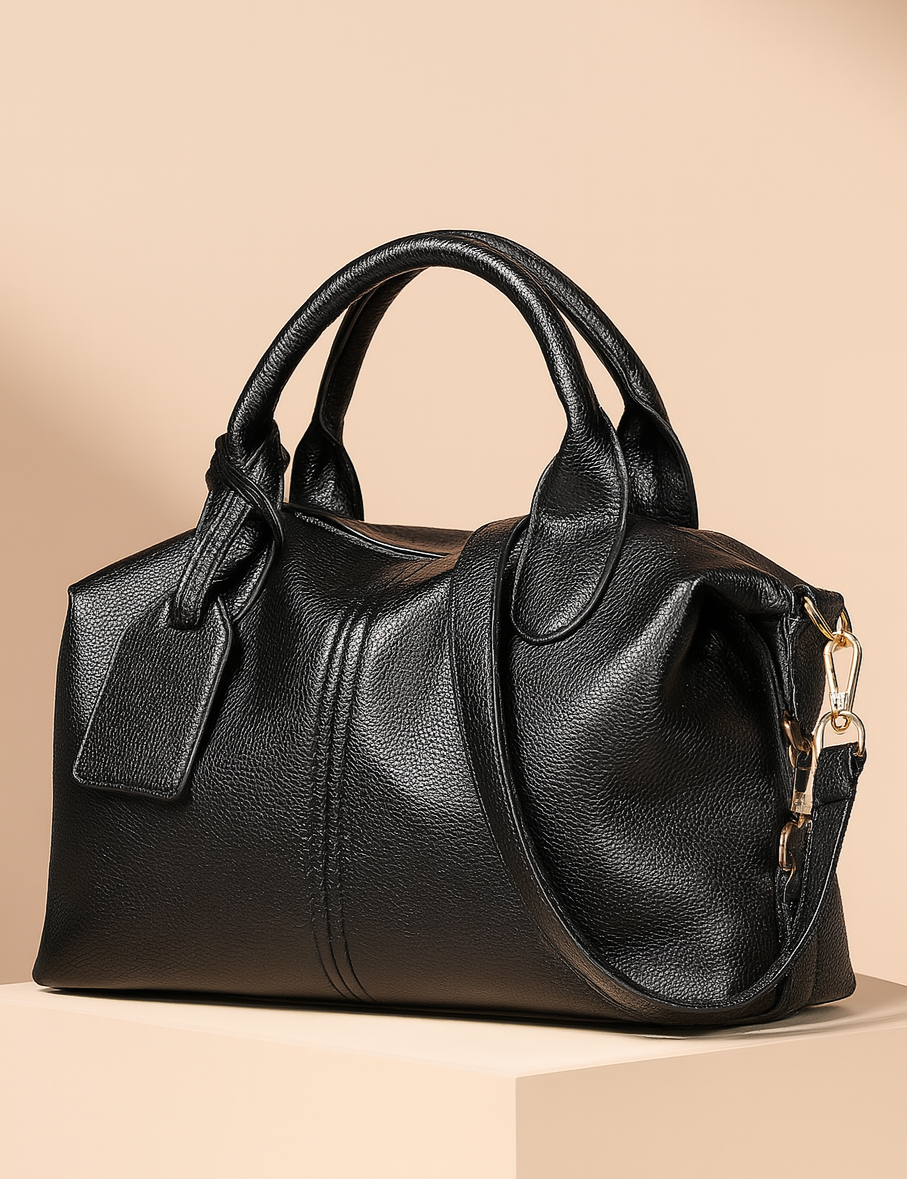 Black pebbled leather handbag with rounded handles, a side tag, and gold hardware, styled on a beige pedestal against a warm sand-to-blush gradient background.