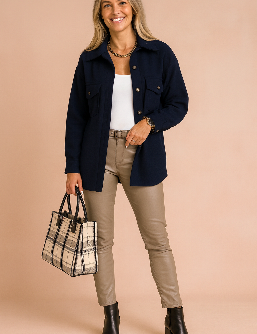 Navy blue button-up shirt jacket with front flap pockets, paired with a white tank top, beige fitted leather-look pants, black ankle boots, and a plaid structured handbag.