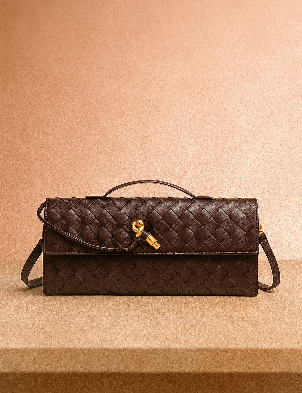 Dark brown woven leather handbag with gold knot clasp, top handle, and wrap-around strap, displayed on a neutral table with a softly lit warm blush background.