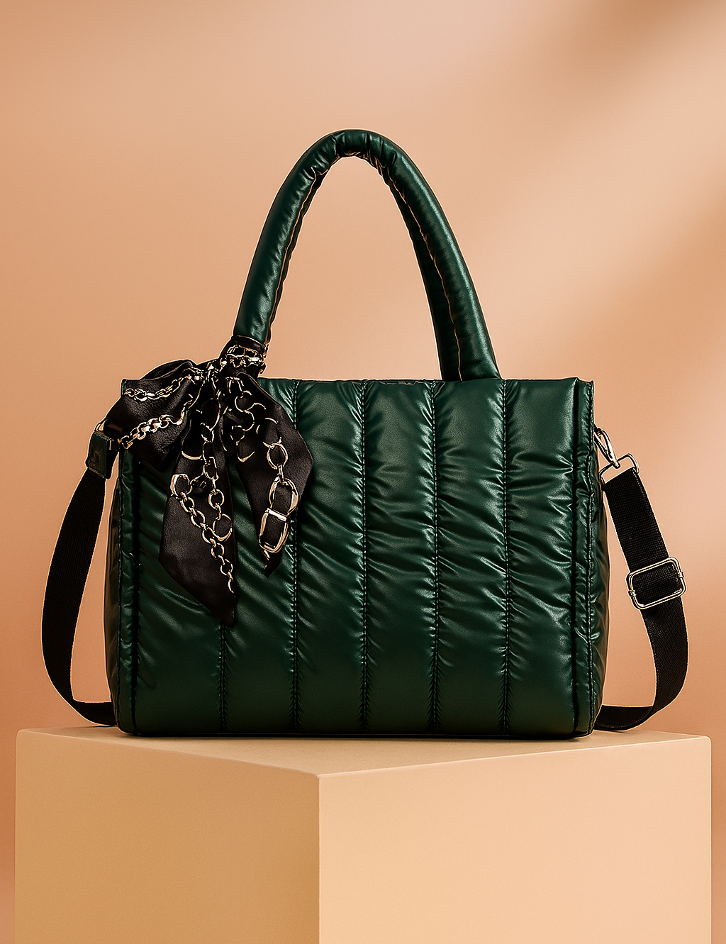 Dark green quilted satin tote bag with top handles, black adjustable strap, and black chain-print scarf tied at the handle, displayed on a pedestal with warm gradient lighting.