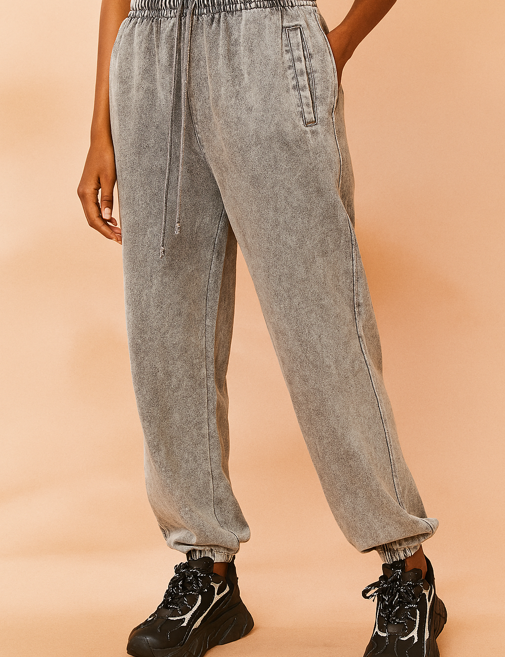 High-waisted gray acid-washed sweatpants with elastic waistband, drawstring tie, side slit pockets, and elasticized cuffs, styled with black chunky sneakers featuring bold white details.