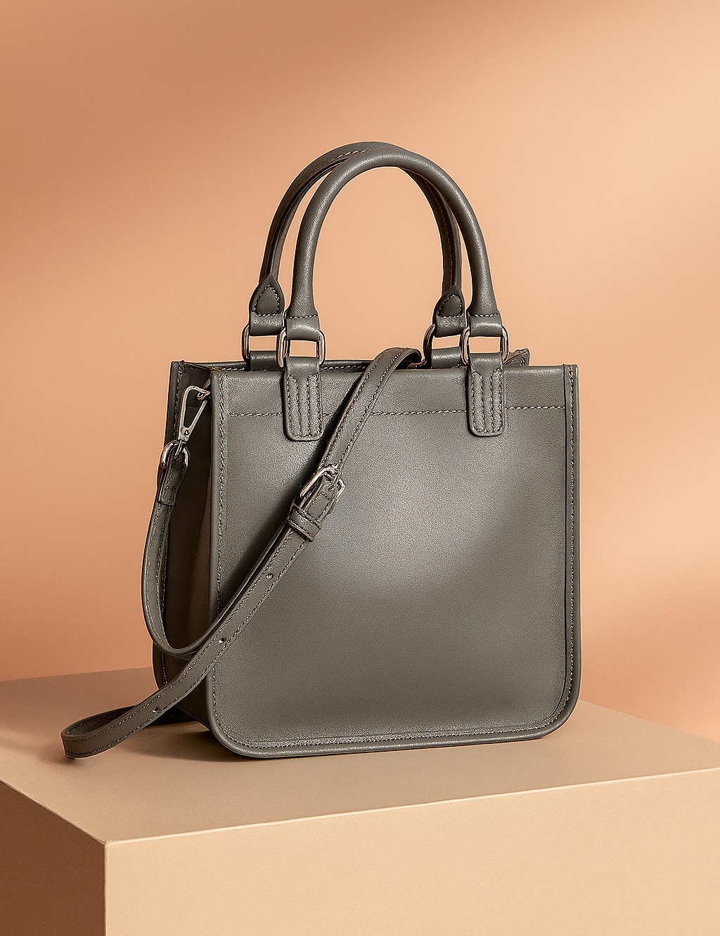Structured gray leather handbag with two top handles, clean edge stitching, and a matching long shoulder strap with silver-tone hardware, presented against a soft warm-toned background.