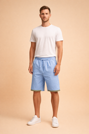 Light blue drawstring summer shorts with elastic waist and geometric hem trim paired with a clean white short sleeve t-shirt and minimalist white low top sneakers, styled for a relaxed European warm weather look