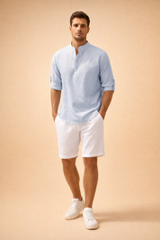 Light blue fine-striped seersucker men’s shirt with band collar and buttoned placket, long sleeves with roll-tab cuffs, paired with clean white tailored shorts and minimalist white low-top leather sneakers, offering a relaxed yet refined warm-weather look.