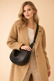 Camel wool coat layered over a neutral beige top and tailored high-waisted trousers, styled with a black leather crescent crossbody bag featuring a wide adjustable strap and polished metal hardware.