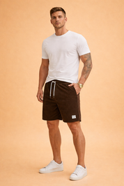 Ribbed corduroy men’s casual shorts in deep brown with elastic waistband, white drawstring, mid-thigh length, straight cut, soft vertical cord texture, and woven logo patch detail.