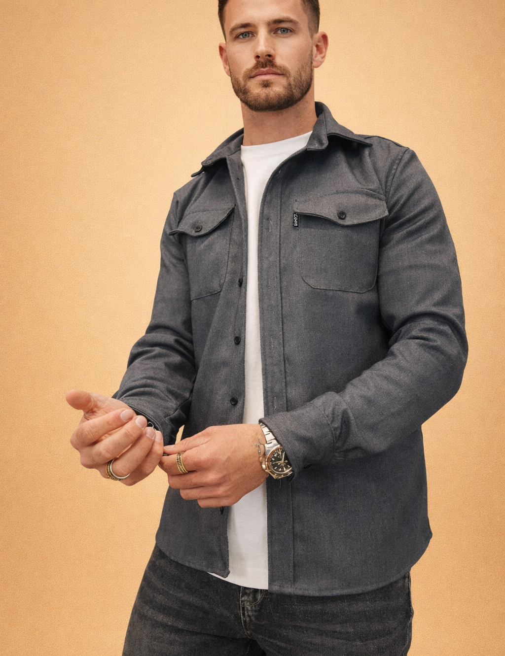 Men’s charcoal grey button-front overshirt with structured collar, dual chest flap pockets, long sleeves with buttoned cuffs, layered over a clean white crew-neck top and styled with slim dark denim trousers