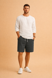 Men’s minimalist summer outfit featuring a clean long-sleeve white shirt paired with tailored ribbed drawstring shorts in a muted cool grey tone, finished with understated neutral footwear for a refined casual look.