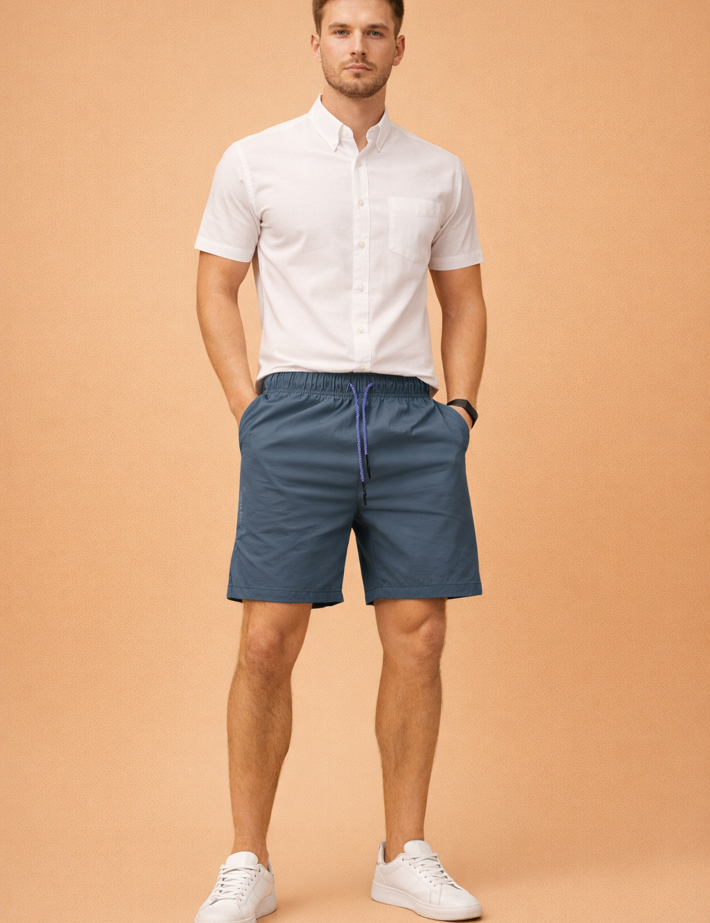 Men’s slate blue lightweight drawstring shorts with elastic waistband and smooth matte finish, paired with a clean white short-sleeve button-up shirt featuring a crisp woven texture, straight hem, and minimal chest pocket detail, styled with classic low-profile white sneakers for a modern summer casual look.