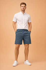 Men’s slate blue lightweight drawstring shorts with elastic waistband and smooth matte finish, paired with a clean white short-sleeve button-up shirt featuring a crisp woven texture, straight hem, and minimal chest pocket detail, styled with classic low-profile white sneakers for a modern summer casual look.