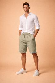 Men’s relaxed summer outfit featuring sage green drawstring shorts with a natural crinkled fabric texture, straight knee-length cut, side pockets, and a subtle geometric trim at the hem, paired with a crisp white long-sleeve button-up shirt worn casually and clean white low-profile sneakers for a modern warm-weather look.