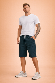 Mid-length men’s casual shorts in a deep teal blue shade, crafted from smooth woven fabric with a clean matte finish, featuring an elastic waistband with white drawstring, straight knee-length cut, minimal seam structure, and a lightweight summer drape paired with a solid short-sleeve crew-neck T-shirt in soft muted teal cotton jersey with a fine, even knit texture.
