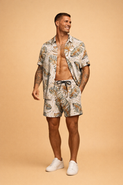 Men’s tropical two-piece summer set featuring a short-sleeve button shirt and matching drawstring shorts with an abstract palm leaf print in beige, olive green, and warm brown tones, paired with clean minimalist low-profile shoes for a polished resort look