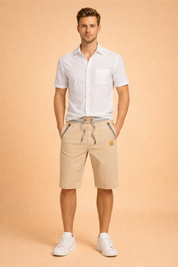 Men’s white short sleeve button-up shirt paired with beige cotton drawstring shorts featuring striped pocket trim, ribbed waistband, and matching beige fabric tone, styled with clean white low-top sneakers for a refined casual summer look.