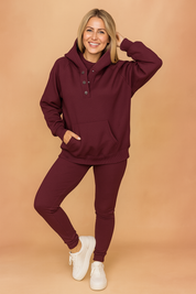 Ribbed burgundy hoodie and matching leggings set featuring a three-button collar and front pouch pocket, styled with clean white sneakers.