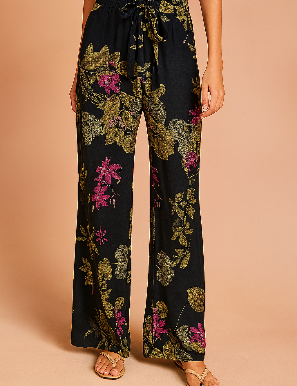 Wide-leg black pants featuring a vivid floral print with olive-green leaves and magenta flowers, styled with a front tie waist and paired with beige sandals.