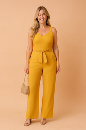 Mustard yellow sleeveless jumpsuit featuring a V-neckline, wide-leg pants, and a center-tied fabric belt, crafted from a lightly textured woven fabric with a clean, solid finish.
