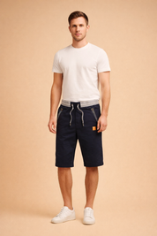 Navy blue men’s knee-length cotton shorts with adjustable drawstring waist, grey ribbed waistband detail, zippered front pockets with striped trim, subtle vertical pinstripe lining at hem, and clean solid fabric finish paired with minimalist white low-top sneakers