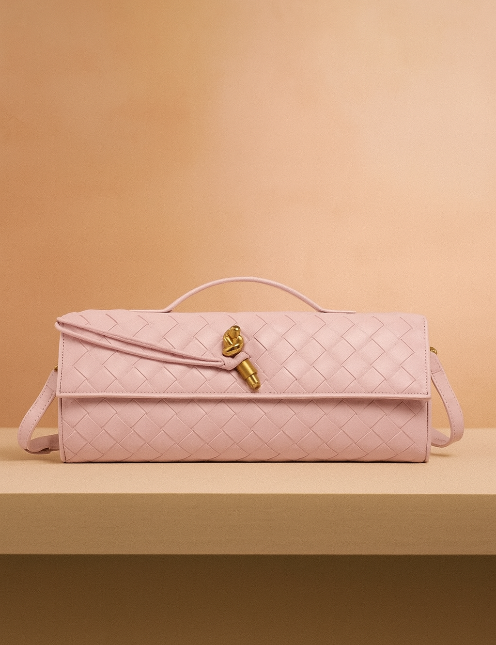 Rectangular pastel pink woven leather handbag with gold knot detail and flap closure, displayed on a beige platform against a warm blush gradient background.