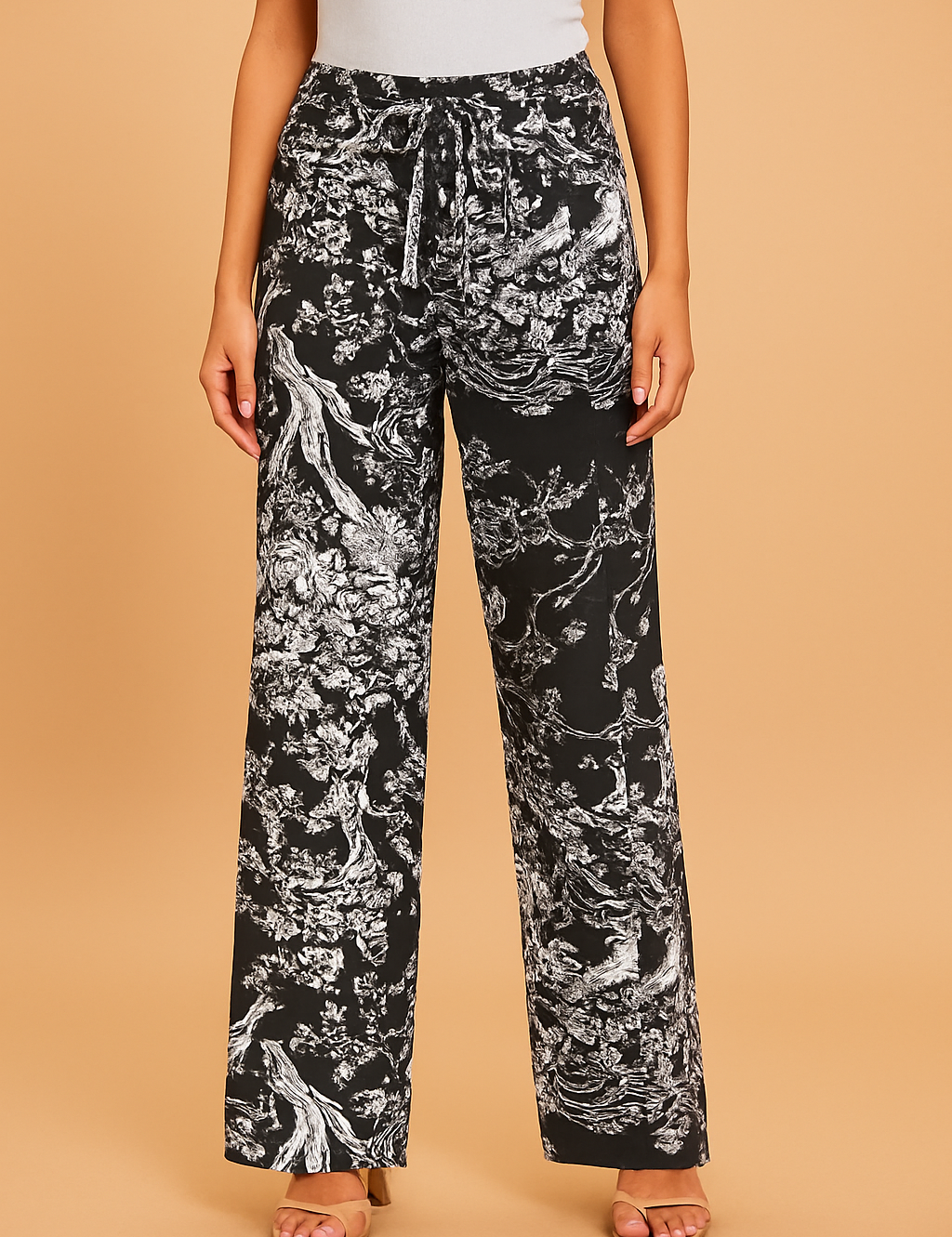 High-waisted black wide-leg pants featuring an intricate white botanical and nature-inspired print with a matching drawstring tie and button, styled with beige strappy sandals.
