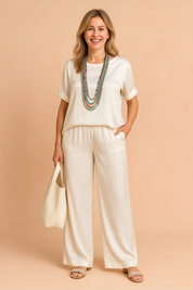 Elegant ivory-white satin co-ord set featuring a loose-fit short-sleeve blouse with rolled cuffs and a soft high-low hemline, paired with wide-leg trousers in matching silky fabric. The set is crafted with a consistent smooth sheen and fluid drape, maintaining the original fabric's light-reflective texture and clean structure.