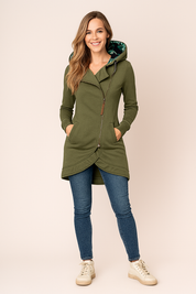 Olive green asymmetrical zip-up hoodie with curved hem, front pockets, and a tropical leaf-print hood lining, styled with slim blue jeans and cream high-top sneakers.