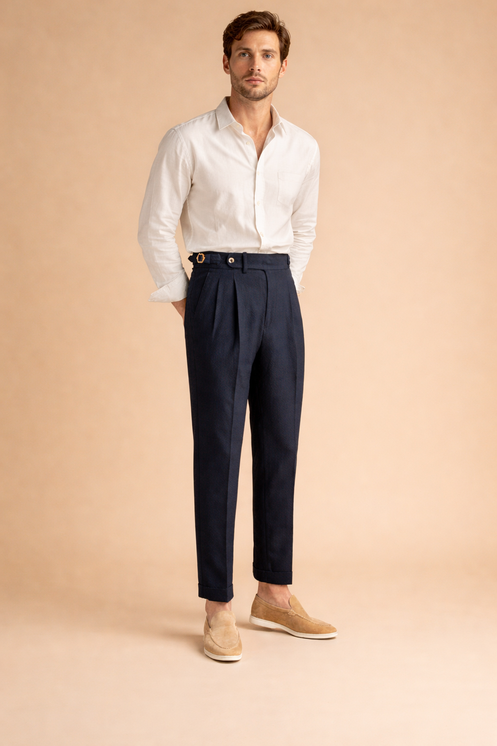Tailored high-waist pleated trousers with a structured waistband, front button closure, belt side adjusters, clean tapered leg, and cuffed hem styled with a long-sleeve button-up shirt and minimalist loafers.