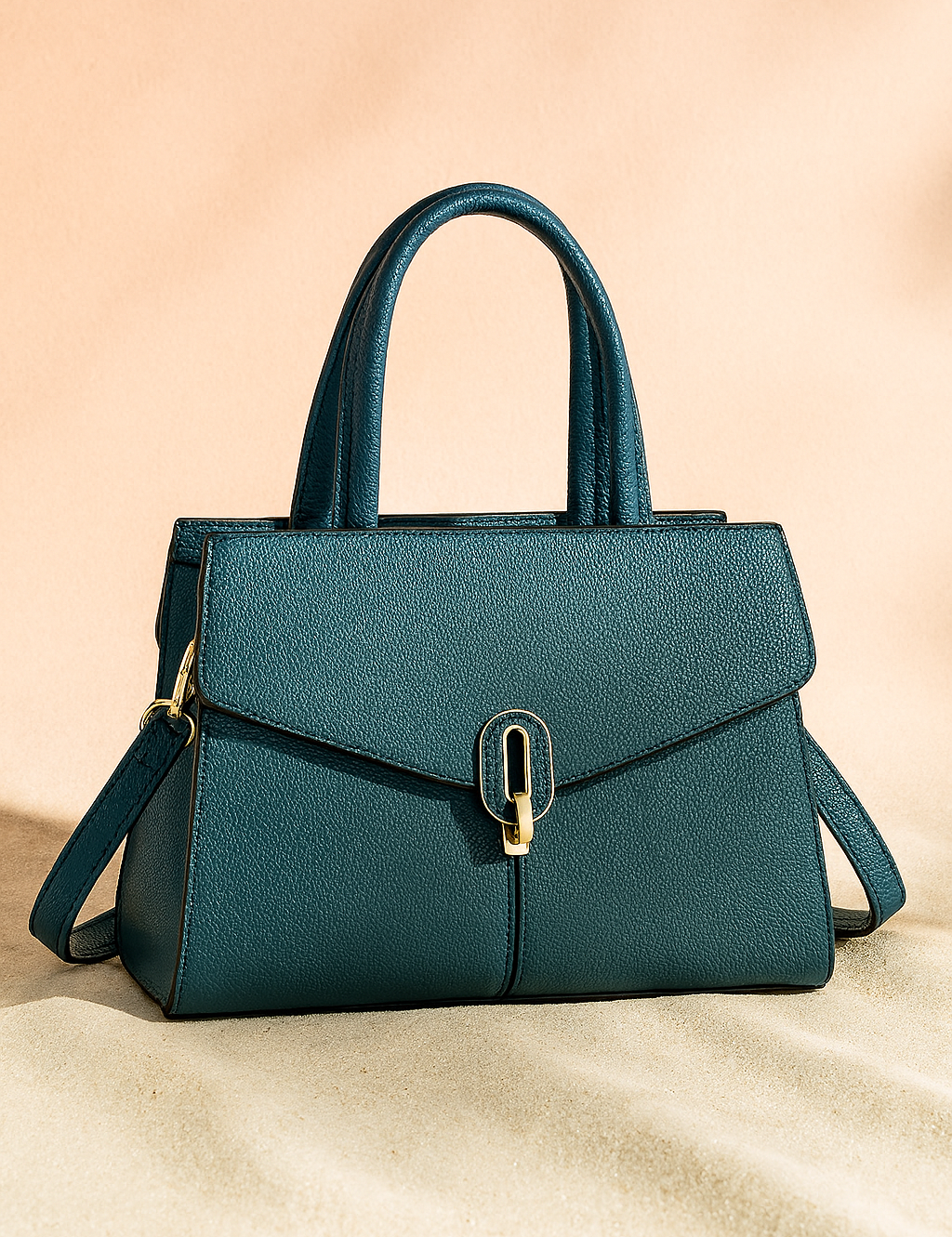Teal pebbled leather handbag with gold-tone clasp, structured top handles, and detachable shoulder strap, shown on a soft sand base and warm pastel background.