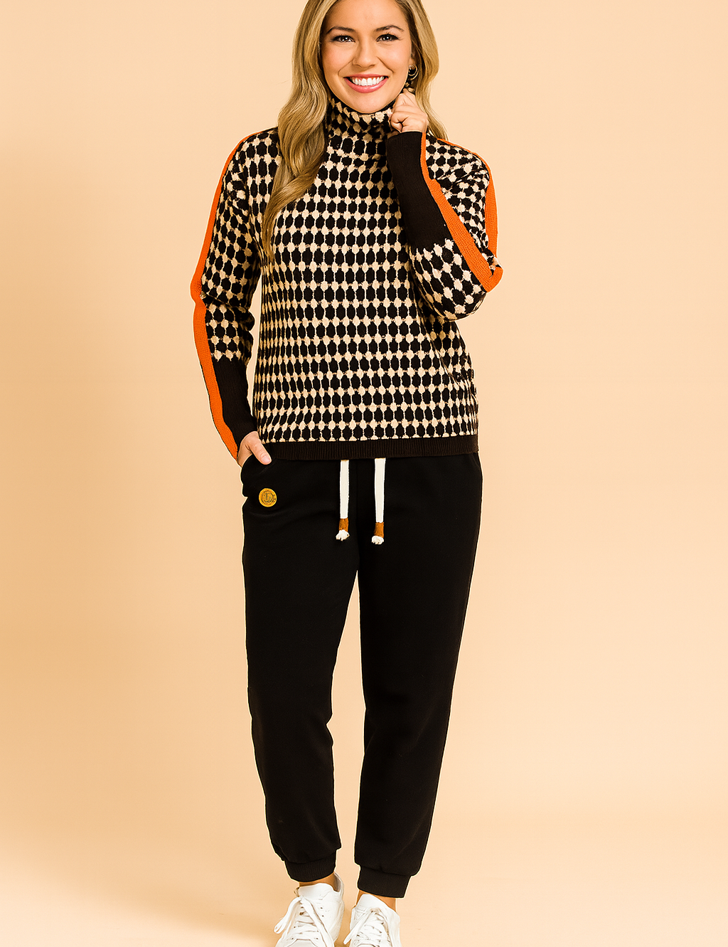 Oversized houndstooth turtleneck sweater with bold orange stripe detailing on sleeves, paired with black jogger pants and white sneakers.