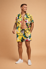 Yellow tropical print short-sleeve button-up shirt with matching drawstring shorts featuring bold floral and leaf motifs in green, white, pink, and black, styled as a coordinated summer set with a relaxed tailored fit and lightweight fabric drape, paired with clean minimalist footwear for a polished resort look